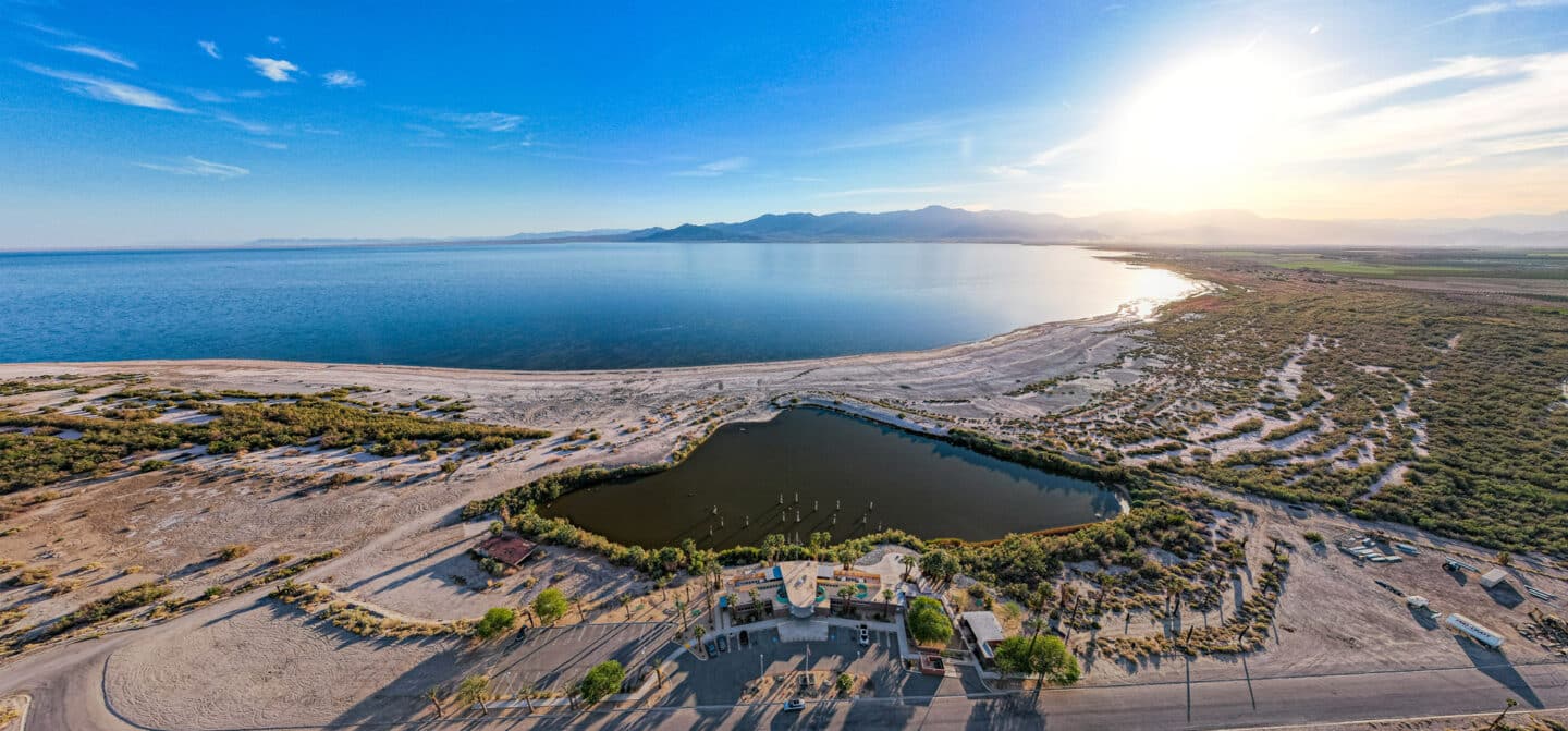 LBL_Top_1995x930px_Salton-Sea_DJI_0946-1440×671 Aerial view of the Salton Sea in Southern California at sunrise, with a visitor center and small lagoon in the foreground and vast desert landscape stretching to distant mountains. This region is a focus of Lawrence Berkeley National Laboratory's geothermal and critical minerals research, including a major study estimating over 3,400 kilotons of lithium in geothermal brines—enough to support large-scale domestic battery production.