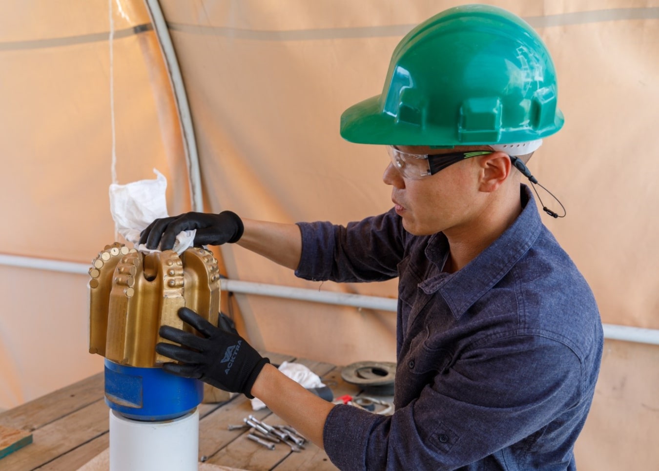 Sandia PDC Jiann Su, Distinguished Member of the Technical Staff at Sandia National Laboratories, inspects and cleans a polycrystalline diamond compact (PDC) drill bit. Wearing safety gear including a green hard hat and protective gloves, Su works at a bench inside a field tent as part of DOE-supported research to improve geothermal drilling performance using data from the Utah FORGE site.