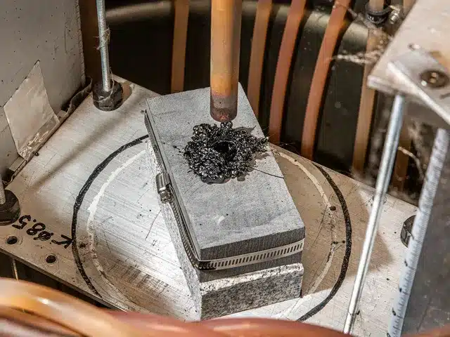 Crane-Geothermal_Final-9093 Close-up view of a laboratory geothermal drilling experiment, showing a heat source melting a hole into a block of granite. The setup includes a metal clamp securing the granite, with black molten residue at the contact point. Tubing and equipment surround the experiment, indicating a controlled testing environment for advanced drilling technologies.