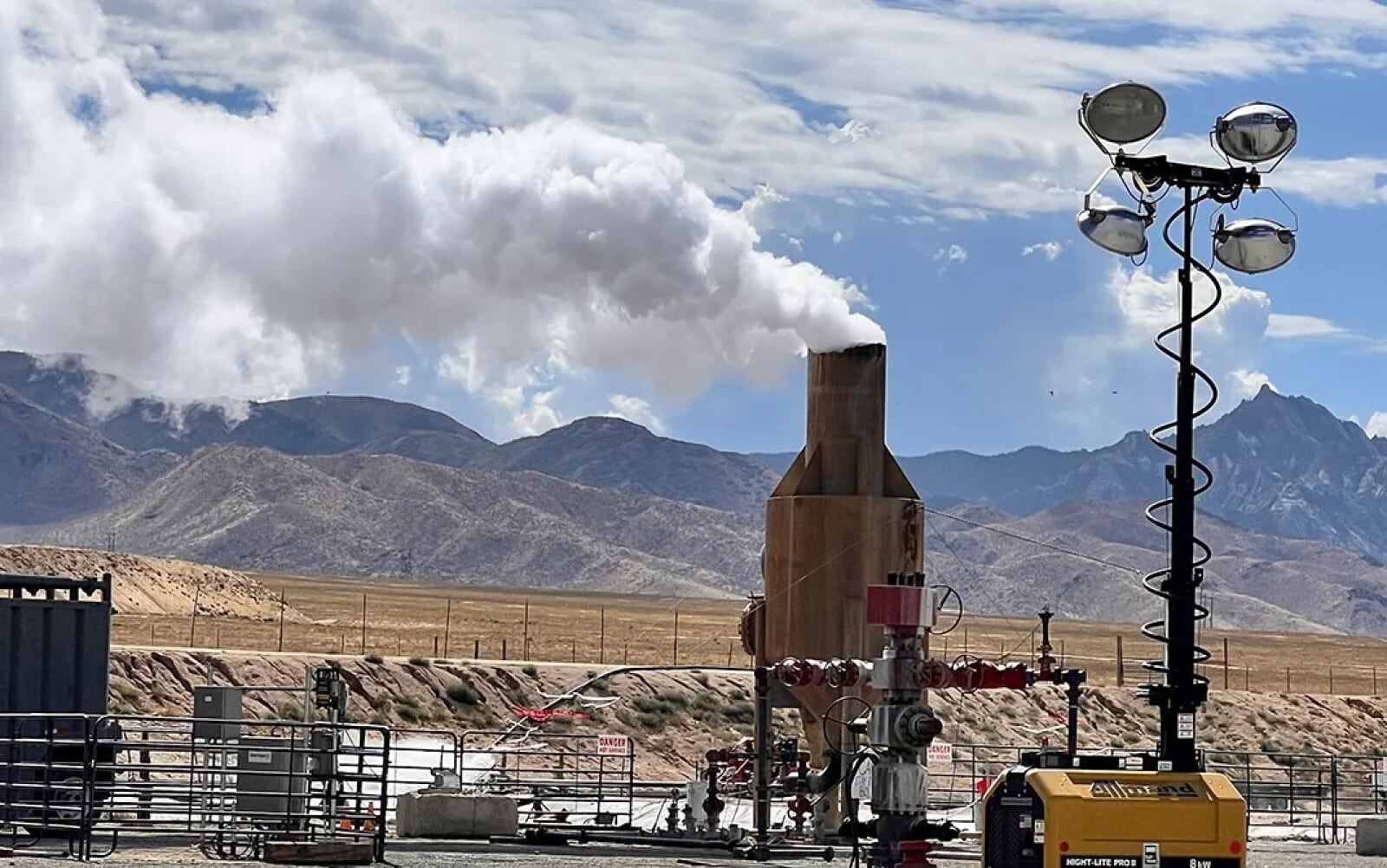 jpt-article Steam rises from a wellhead at the Utah FORGE geothermal research site in Milford, Utah, with rugged desert mountains in the background. The image shows geothermal infrastructure including a wellhead, piping system, and steam separator, along with a portable light tower labeled “NIGHT-LITE PRO II.” The scene is set against a partly cloudy sky with a view of the Mineral Mountains, highlighting the remote and industrial nature of this advanced geothermal energy project.