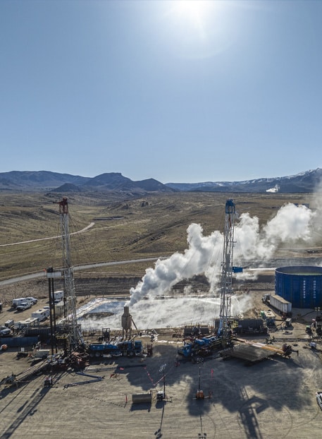 Aerial view of the Utah FORGE geothermal research site near Milford, Utah, showing two drilling rigs, rising steam from well testing, support vehicles, and a large blue water tank, set against a backdrop of mountains under a bright sun.