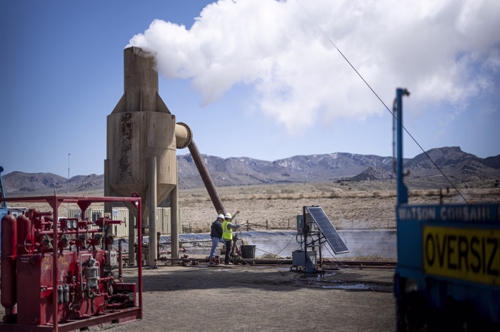 Workers stand near a geothermal wellhead releasing steam at the Utah FORGE site in Milford, Utah. Surrounded by drilling equipment, a solar panel, and mountain landscape, the image captures ongoing enhanced geothermal energy research.
